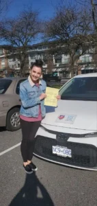 A smiling woman in a denim jacket and black pants holds a yellow form beside the front of a white car.