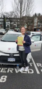 A smiling woman in a navy jacket and black leggings holds a yellow form in front of a white car with the driver door open.