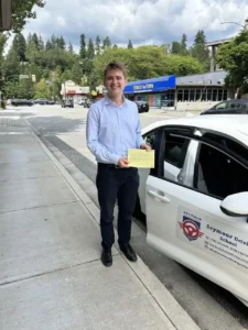 A smiling young man in a light blue button-down shirt and black pants holds a yellow form beside the open door of a white Seymour Driving School car on a sidewalk.