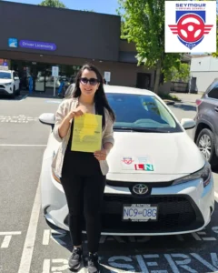A woman in a beige blazer, black clothes, and sunglasses holds a yellow form in front of a white Seymour Driving School car outside a driver licensing office.