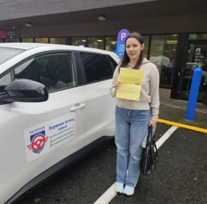 A woman in a beige hoodie and light blue jeans holds a yellow form and a black bag beside a white Seymour Driving School car.