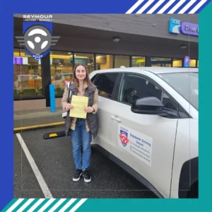 A woman in a beige jacket, dark vest, and blue jeans smiles while holding a yellow form beside a white Seymour Driving School car in a parking lot.