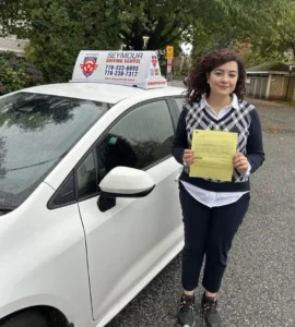 A woman in a black-and-white argyle vest and navy pants holds a yellow form beside a white Seymour Driving School car.