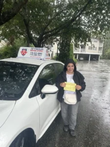 A woman in a black hooded jacket, white top, and grey pants holds a yellow form beside a white Seymour Driving School car on a wet street under trees.