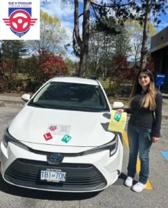 A woman in a black jacket and blue jeans holds a yellow form with a green N beside a white Seymour Driving School car.