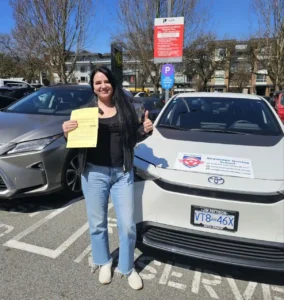 A woman in a black jacket and light blue jeans holds a yellow form and gives a thumbs-up beside a white Seymour Driving School car.