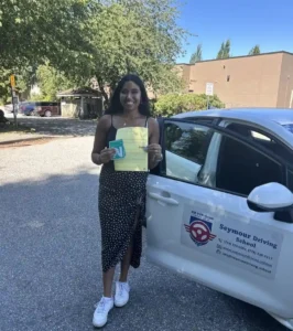 A woman in a black polka-dot dress and white sneakers holds a yellow form and a green N sign beside the open door of a white Seymour Driving School car.