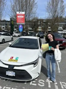 A woman in a black t-shirt and blue jeans holds a yellow form and a green N sign beside a white Seymour Driving School student-driver car, with a white jacket over her arm.
