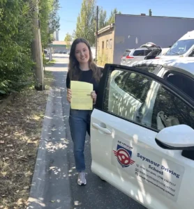 A woman in a black T-shirt and blue jeans holds a yellow form beside the open door of a white Seymour Driving School car.