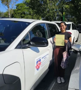 A woman in a black T-shirt and brown trousers holds a yellow form and a patterned tote bag beside a white Seymour Driving School car under leafy trees.