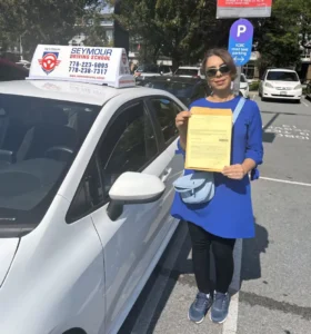 A woman in a blue dress, black leggings, and sunglasses holds a yellow form beside a white Seymour Driving School car in a parking lot.