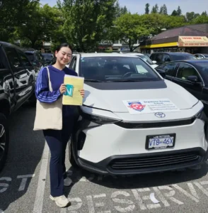 A woman in a blue striped long-sleeve shirt and dark pants holds a yellow form and a green N sign beside a white Seymour Driving School car.