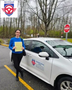 A woman in a blue sweater and black pants holds a yellow form beside a white Seymour Driving School car.