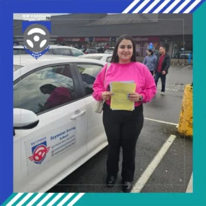 A woman in a bright pink sweater and black pants smiles while holding yellow forms beside a white Seymour Driving School car in a parking lot.
