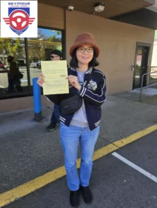 A woman in a brown bucket hat, navy varsity jacket, and blue jeans holds a yellow form on a sidewalk outside a building.