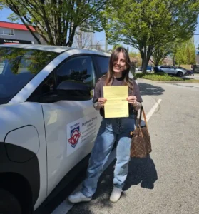 A woman in a brown t-shirt and light blue jeans holds a yellow form beside a white Seymour Driving School car on a sunny street.