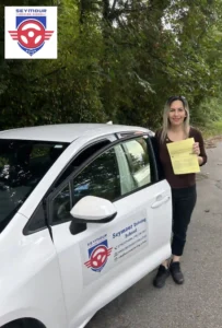 A woman in a brown top and black pants holds a yellow form beside the open door of a white Seymour Driving School car.