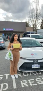 A woman in a brown tracksuit holds a yellow form in front of a white Seymour Driving School car.