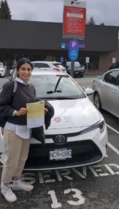 A woman in a dark grey sweater and beige pants smiles while holding a yellow form and a black jacket in front of a white car in a reserved parking stall.