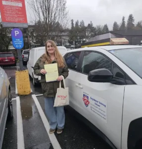 A woman in a green jacket holds a yellow form beside a white Seymour Driving School car. An ICBC road-test parking sign is beside her.