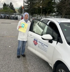 A woman in a light blue hijab, white hoodie, and blue jeans holds a yellow form and a green N sign beside the open door of a white Seymour Driving School car in a parking lot.