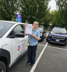 A woman in a light blue shirt and dark jeans holds up a white printed form beside a white Seymour Driving School car in a parking lot.