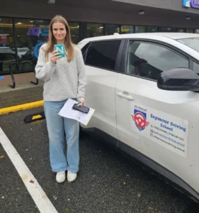 A woman in a light grey sweater and light blue jeans holds a green N sign, a black phone, and papers beside a white Seymour Driving School car.