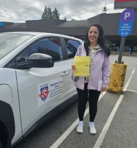 A woman in a lilac jacket over a floral top, black pants, and white sneakers holds a yellow form beside a white Seymour Driving School car near an ICBC road-test parking sign.