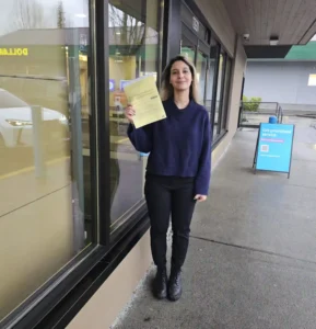 A woman in a navy sweater and black pants holds a yellow form on the sidewalk outside a glass storefront.
