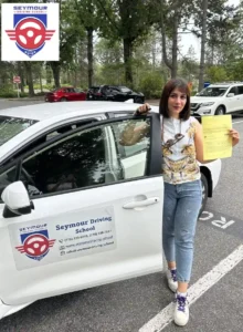 A woman in a patterned sleeveless top and light jeans holds a yellow form beside the open door of a white Seymour Driving School car.