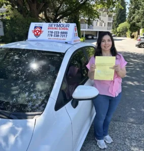 A woman in a pink top and blue jeans holds a yellow form beside a white Seymour Driving School car with a roof sign.