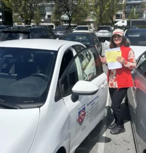 A woman in a red vest and a patterned shirt holds a yellow form beside the open door of a white Seymour Driving School car.