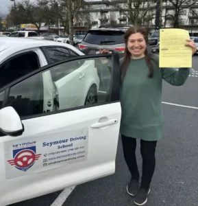A woman in a sage-green sweatshirt and black pants holds up a yellow form beside the open door of a white Seymour Driving School car.