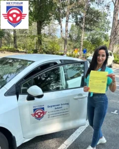 A woman in a teal blouse and blue jeans holds a yellow form beside the open door of a white Seymour Driving School car, with the school logo overlaid in the corner.