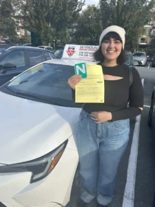 A woman in a white beanie, a black cutout top, and blue jeans holds a yellow form and a green N sign beside a white Seymour Driving School car.