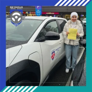 A woman in a white beanie, cream puffer jacket, and light jeans smiles while holding a yellow form beside a white Seymour Driving School car on a wet lot.