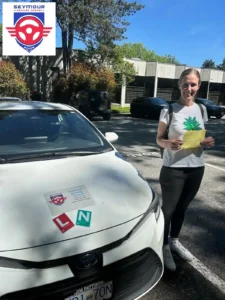 A woman in a white graphic t-shirt and dark pants holds a yellow form beside the front of a white Seymour Driving School car.