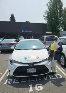 A woman in a white hijab, sunglasses, navy jacket, and light pants holds a yellow form beside a white car with green N and red L signs on the hood outside a driver licensing office.
