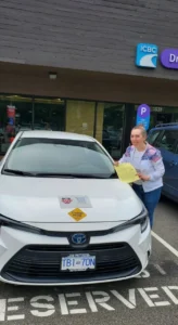 A woman in a white jacket with pink and blue sleeves and blue jeans holds a yellow form beside a white car with a yellow student-driver sign outside an ICBC office.