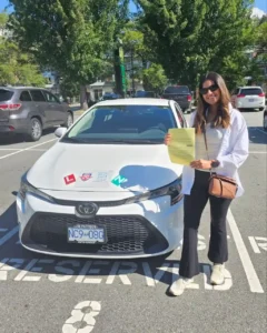 A woman in a white shirt, black pants, and sunglasses holds a yellow form in front of a white Seymour Driving School car with a red L sticker and a green N sticker in a sunny parking lot.