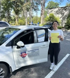 A woman in a white T-shirt and black leggings holds a yellow form beside the open door of a white Seymour Driving School car.