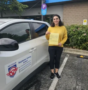 A woman in a yellow sweater and black pants holds a yellow form beside a white Seymour Driving School car.