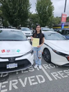 A woman in ripped jeans holds a yellow form between two white cars in a road-test parking lot.