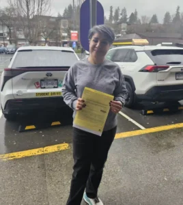 A woman with short grey hair, a grey sweatshirt, and black pants holds a yellow form in front of the rear of a white student-driver car.