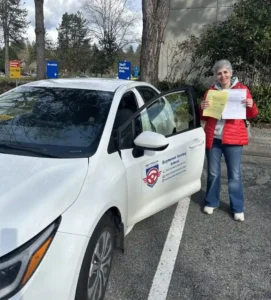 A woman with short grey hair, wearing a red puffer jacket and blue jeans, holds a yellow form and a white sheet beside the open door of a white Seymour Driving School car.