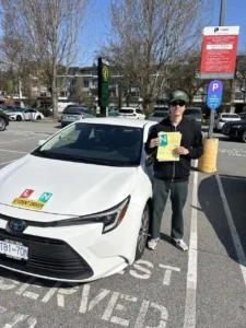 A young man in a black hoodie, green cap, and sunglasses holds a yellow form and a green N sign beside a white Seymour Driving School student-driver car.