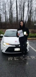 A young man in a black hoodie holds a white paper and a phone in front of a white car on a wet parking lot.