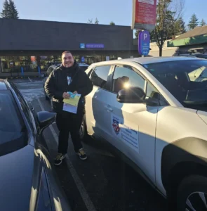 A young man in a black jacket holds a yellow form and a green N sign beside a white Seymour Driving School car outside a Driver Licensing office.