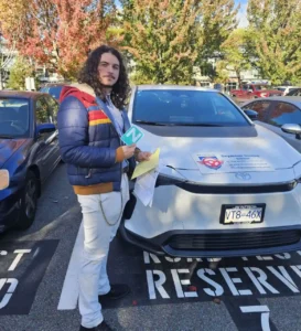 A young man in a navy puffer jacket with red and yellow stripes and white pants holds a green N sign and yellow papers beside a white Seymour Driving School car.