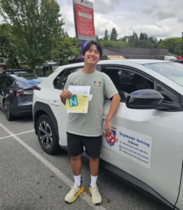 A young man in a sage green T-shirt, black shorts, and white sneakers holds a yellow form and a green N sign beside a white Seymour Driving School car.